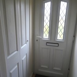 Photo showing a modern white UPVC front door, with two long leaded lattice windows, a white door handle and letterbox, and rectangular door panels, which match the adjacent porch cupboard, where coats and shoes can be hidden away out of sight.  A dark doormat and tiled porch floor can be seen at the base of the front door.